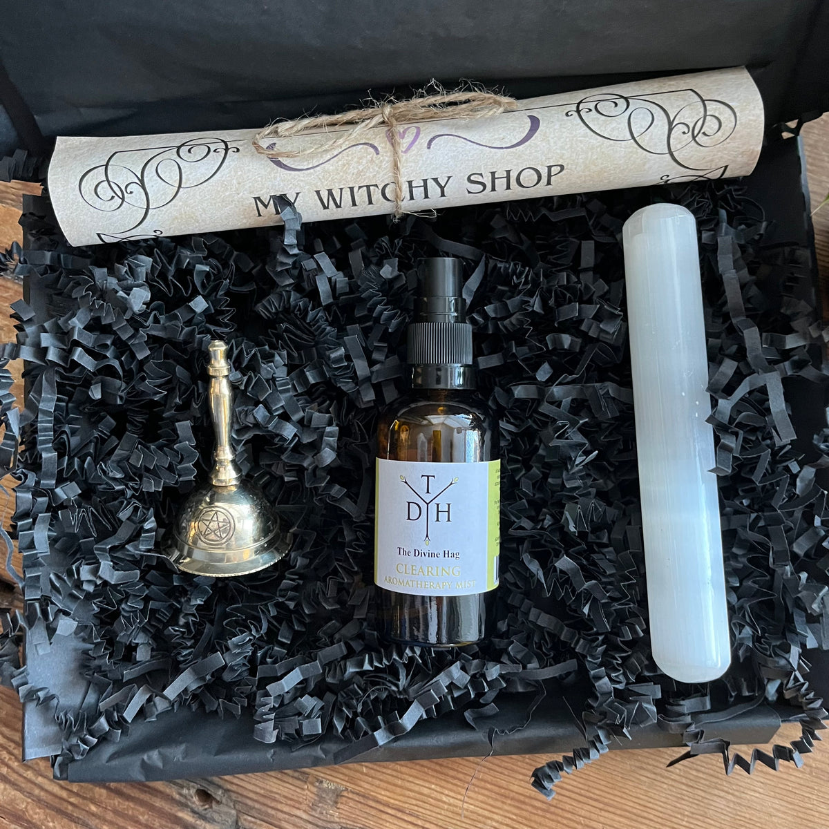 Box with 'My Witchy Shop' scroll, bottle of energy clearing room spray, Selenite wand and bell on black crinkled paper.