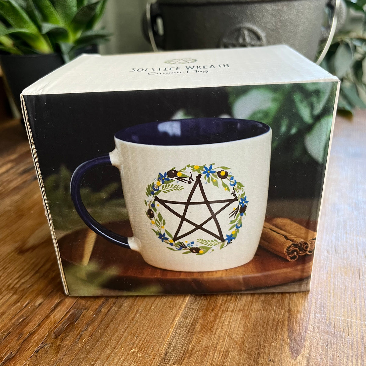 A ceramic mug with a cream exterior and dark blue interior, featuring a black pentagram enclosed in a floral wreath design. The mug sits on a wooden table with its product box visible in the background.