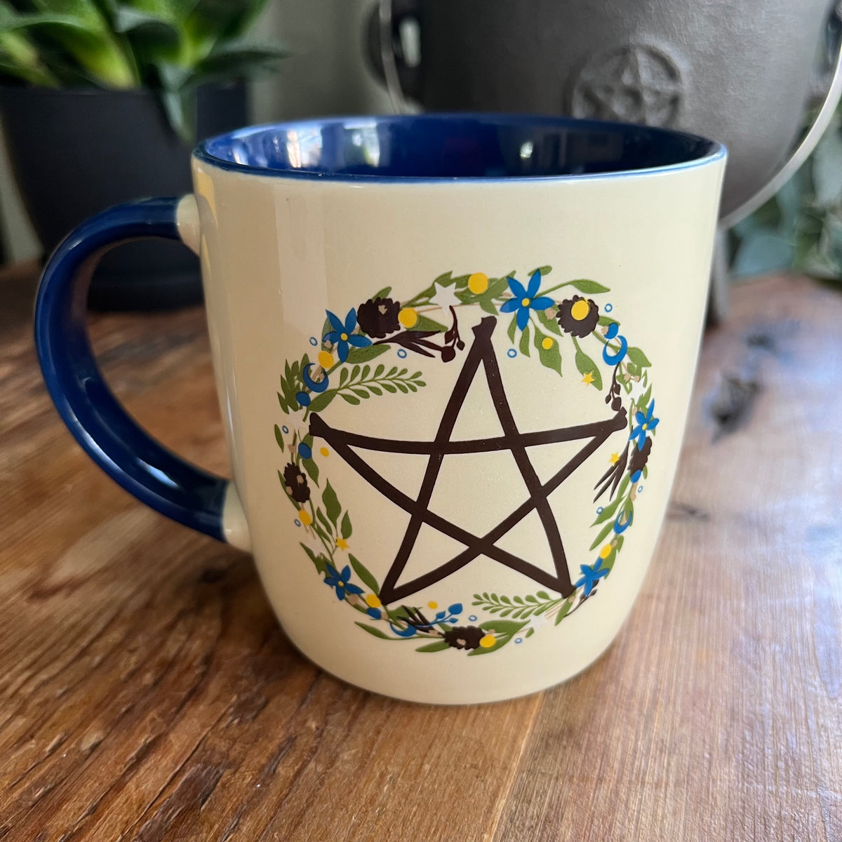 A ceramic mug with a cream exterior and dark blue interior, featuring a black pentagram enclosed in a floral wreath design. The mug sits on a wooden table 