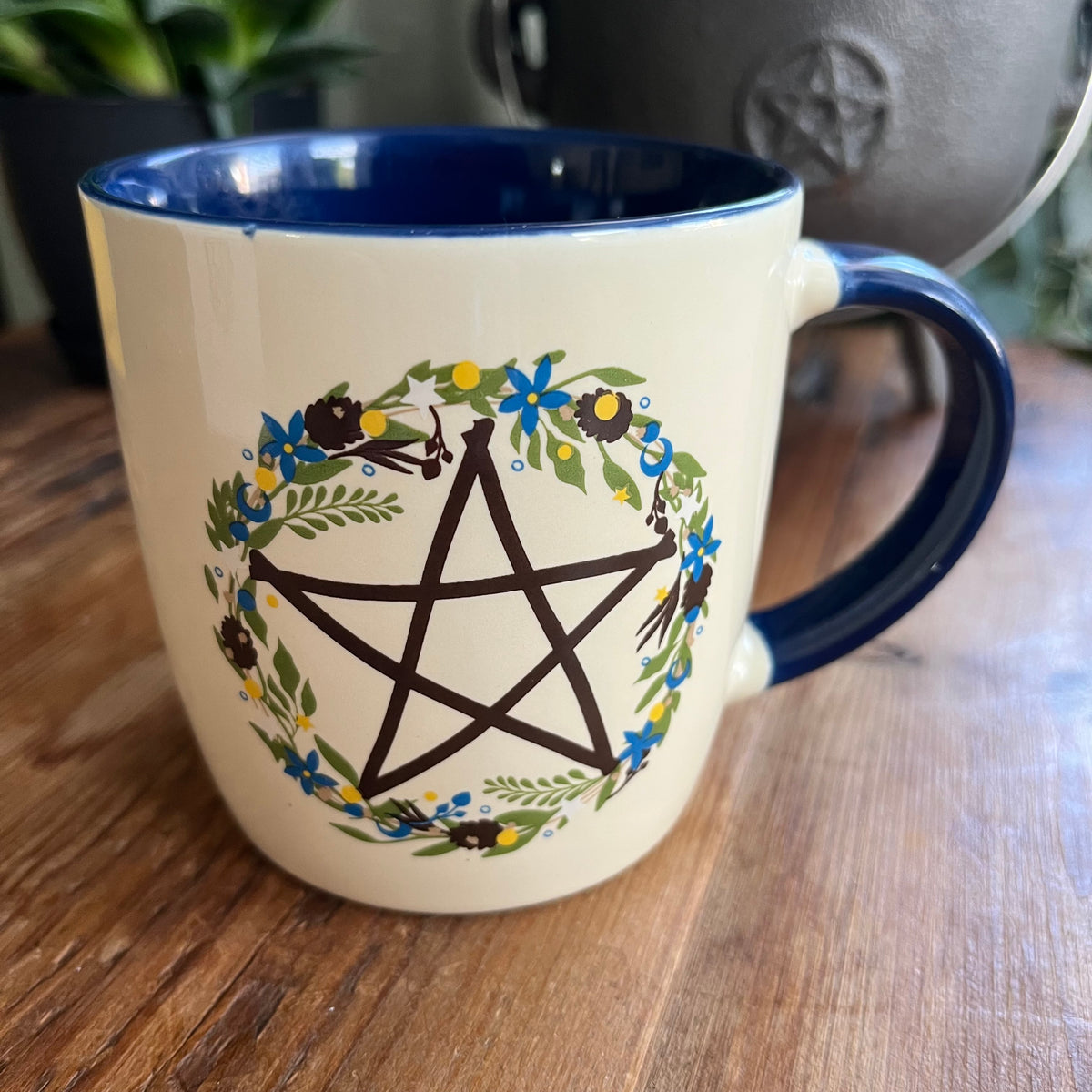 A ceramic mug with a cream exterior and dark blue interior, featuring a black pentagram enclosed in a floral wreath design. The mug sits on a wooden table 