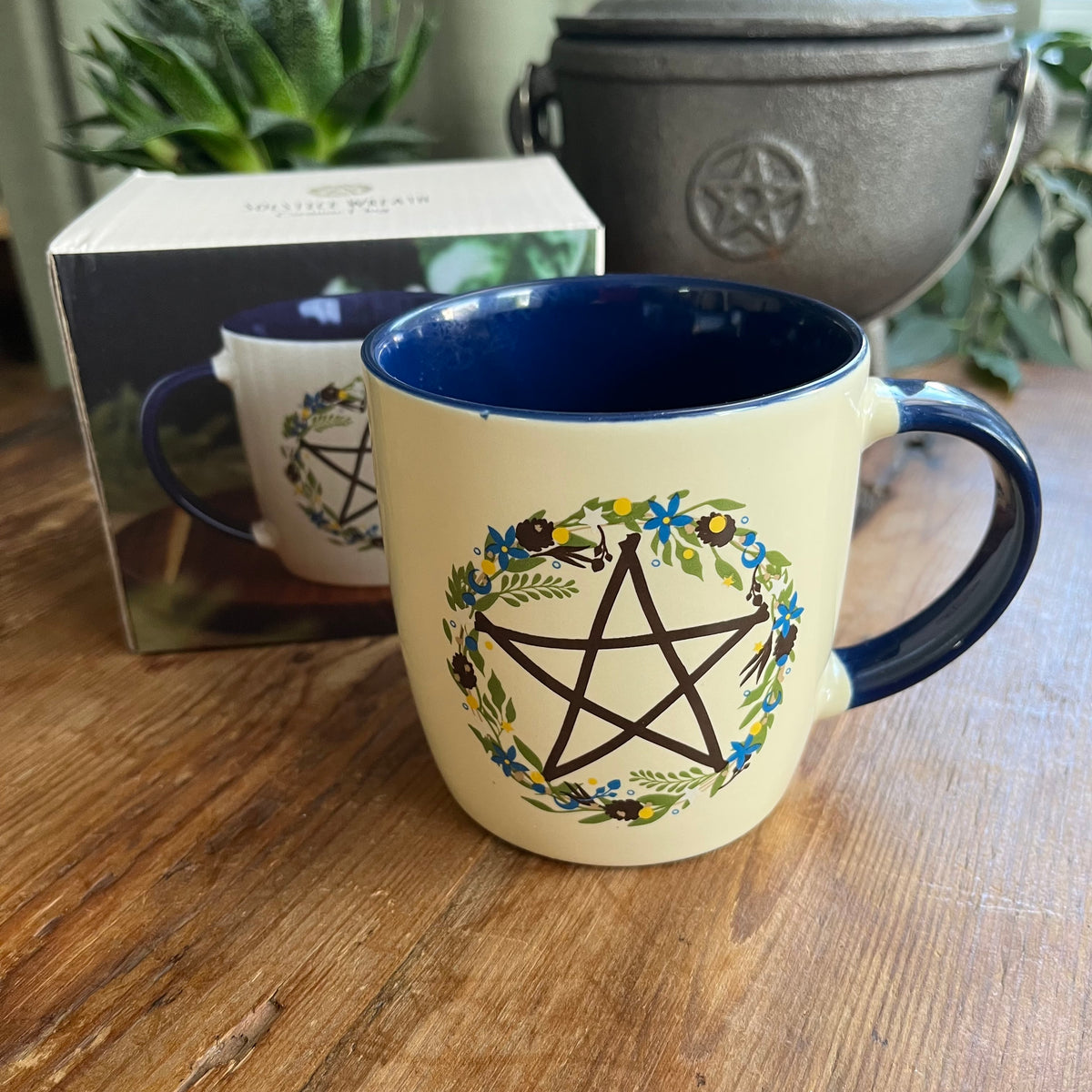 A ceramic mug with a cream exterior and dark blue interior, featuring a black pentagram enclosed in a floral wreath design. The mug sits on a wooden table with its product box visible in the background.