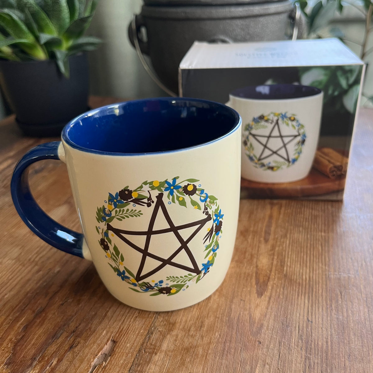 A ceramic mug with a cream exterior and dark blue interior, featuring a black pentagram enclosed in a floral wreath design. The mug sits on a wooden table with its product box visible in the background.