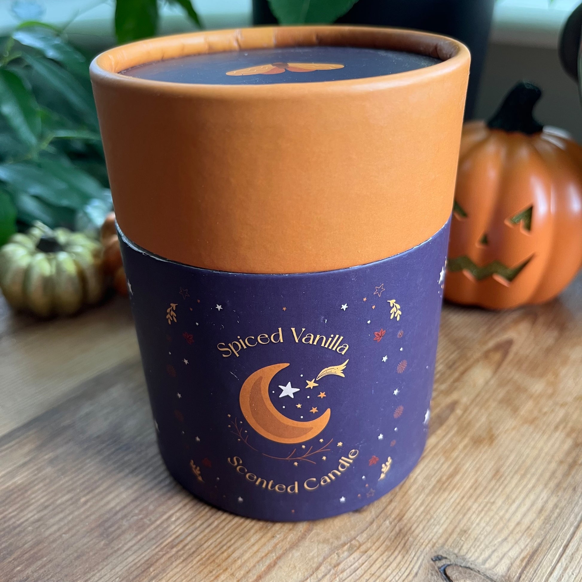 Spiced Vanilla scented candle in a decorative container on a wooden surface with pumpkins in the background.