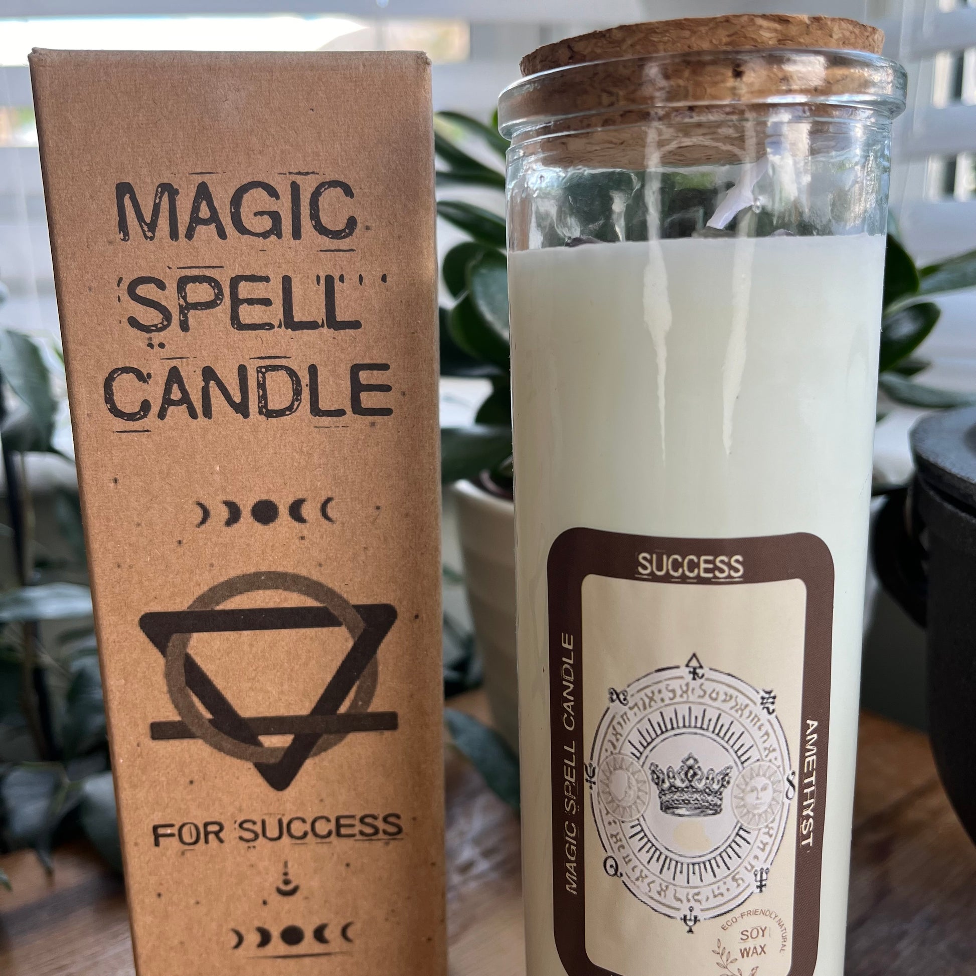 Tall glass spell candle with cork lid. The label on the candle features an image of a crown and the words Success, amethyst and Lavender and Fennel. The candle itself is white with Amethyst gemstones embedded within it. Alongside the glass Magic spell candle is it's brown cardboard box which has the words magic spell candle for Success, soy wax candle with amethyst gemstones and Lavender and Fennel fragrance.