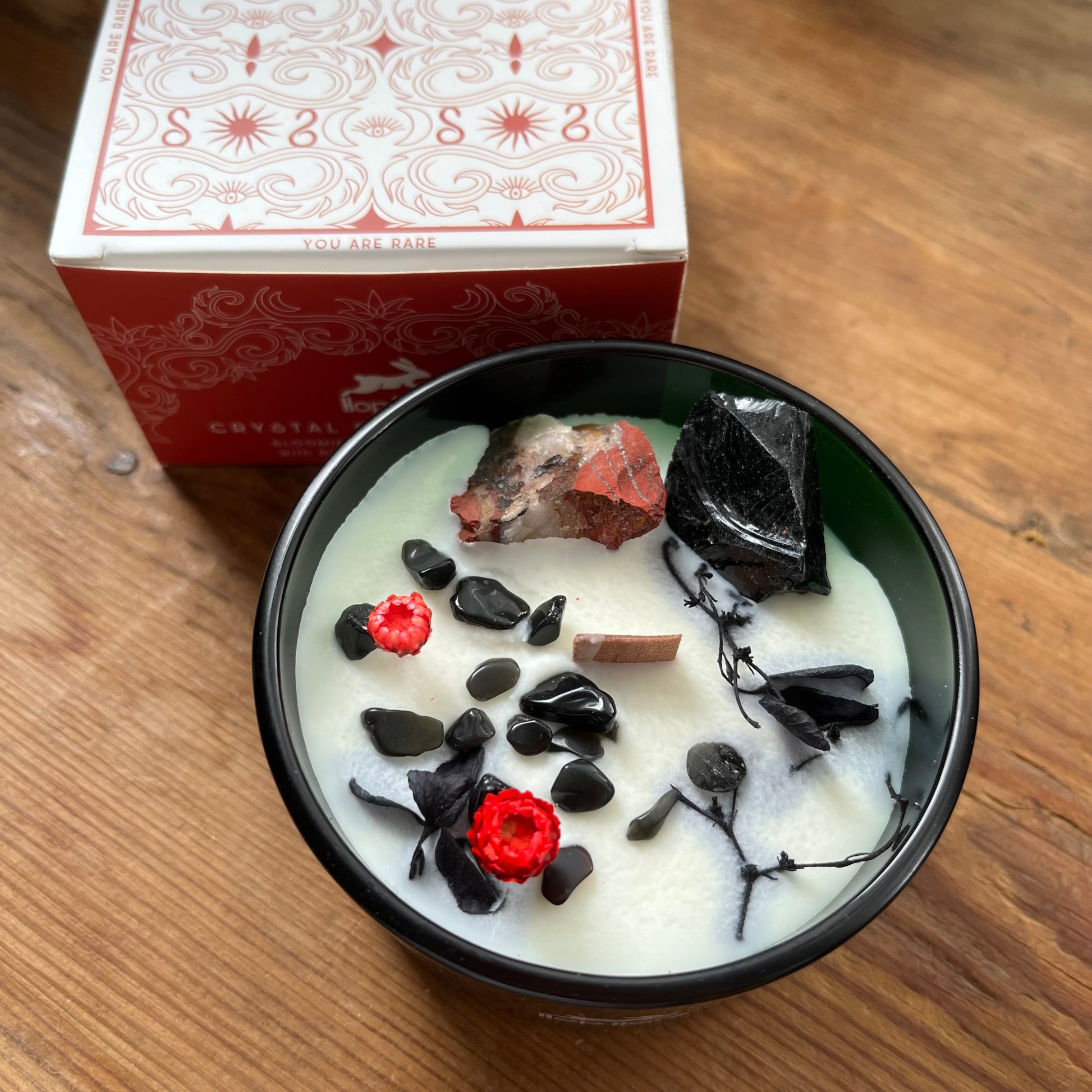 Candle with rocks and crystals in a green container on a wooden surface, with a presentation box in the background.