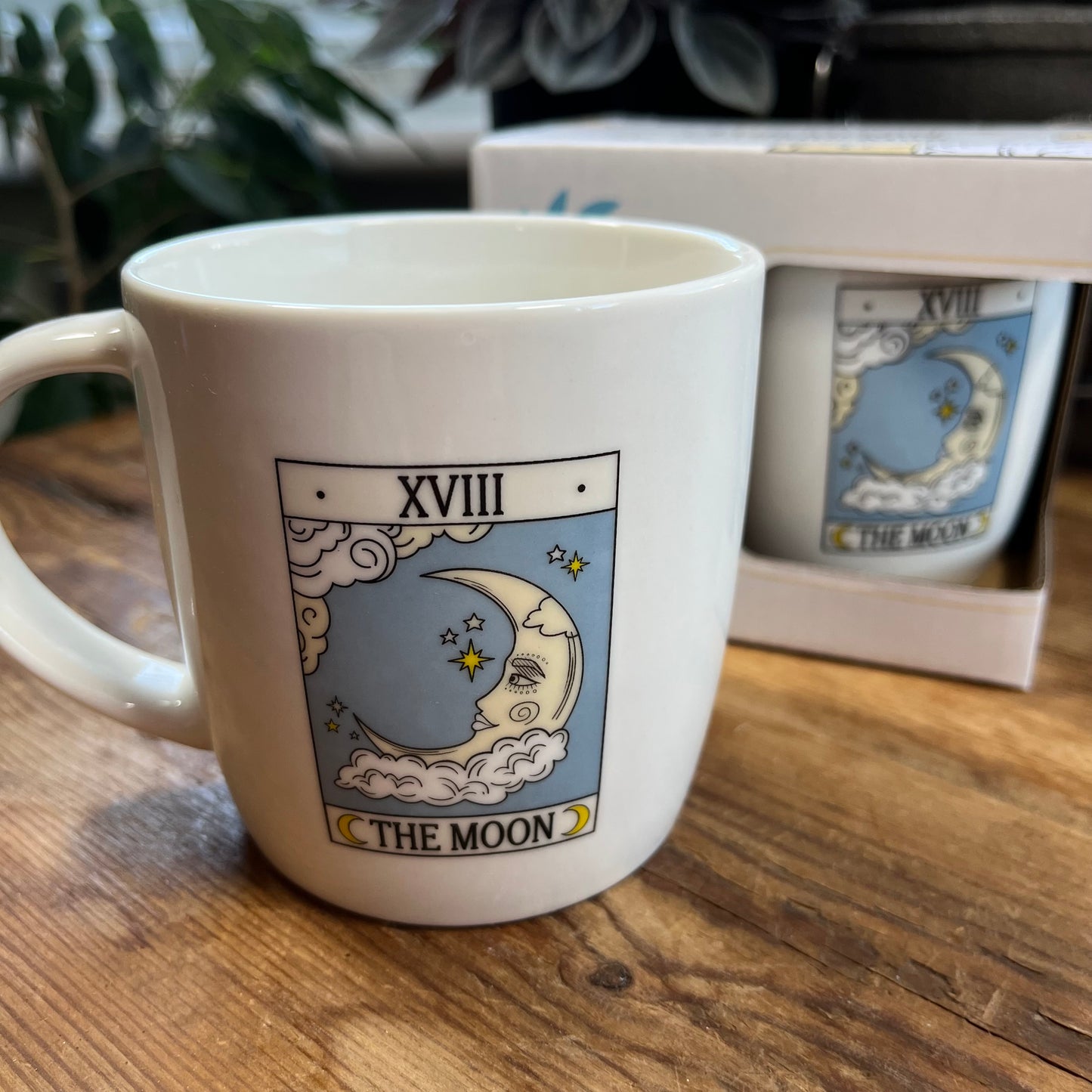 White mug with 'The Moon' tarot card design on a wooden surface, with another mug and box in the background.