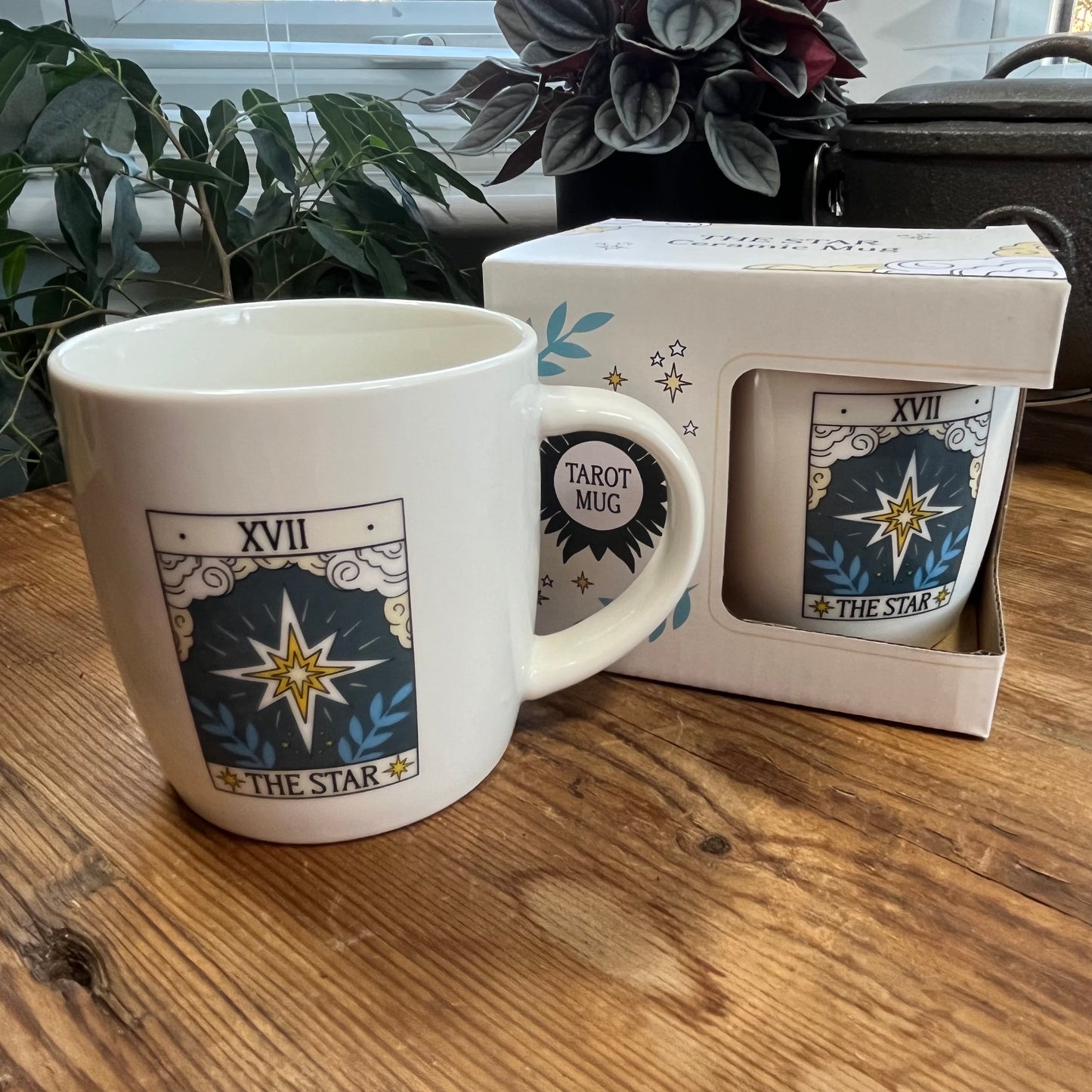 White mug with 'The Star' tarot card design on a wooden surface, next to its packaging.