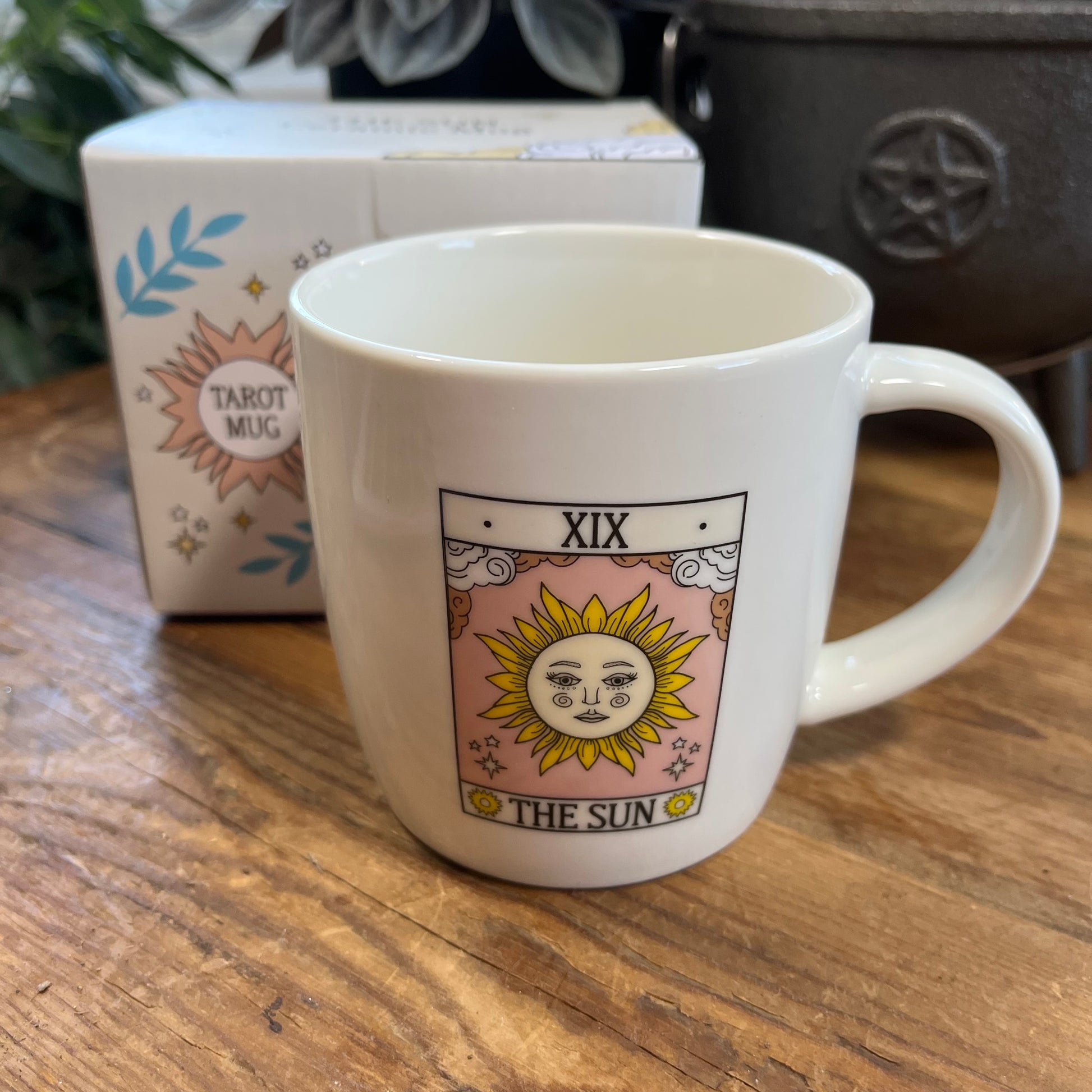 White mug with 'The Sun' tarot card design on a wooden surface, with a box labeled 'Tarot Mug' in the background.