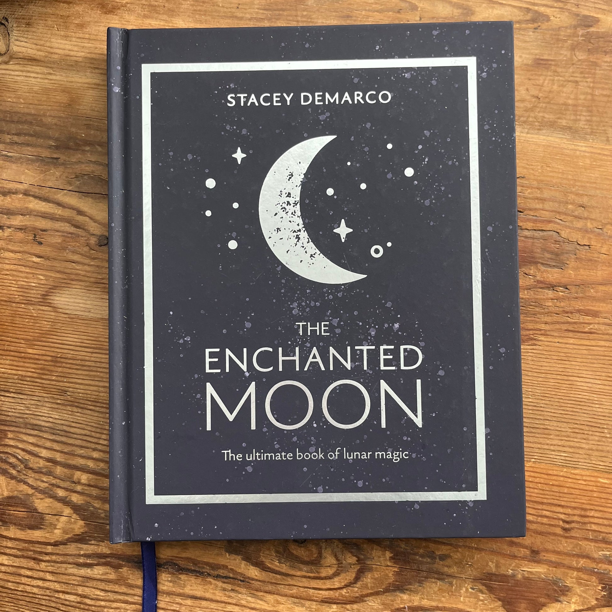 The Enchanted Moon book by Stacey Demarco, featuring a dark slate-gray cover with a white crescent moon and stars, placed on a rustic wooden surface.