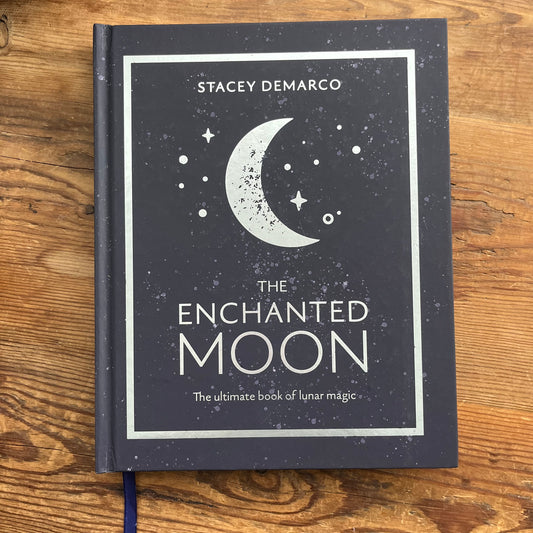The Enchanted Moon book by Stacey Demarco, featuring a dark slate-gray cover with a white crescent moon and stars, placed on a rustic wooden surface.