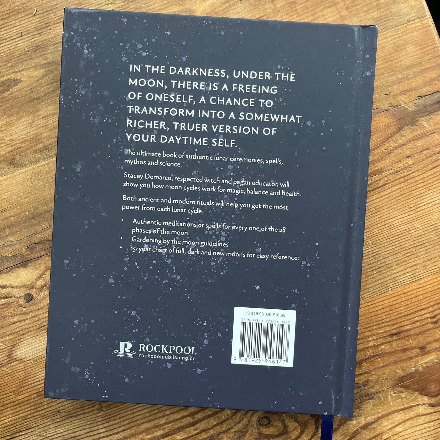 The back cover of a dark blue book lies on a wooden surface. The cover features white text describing lunar ceremonies and spells