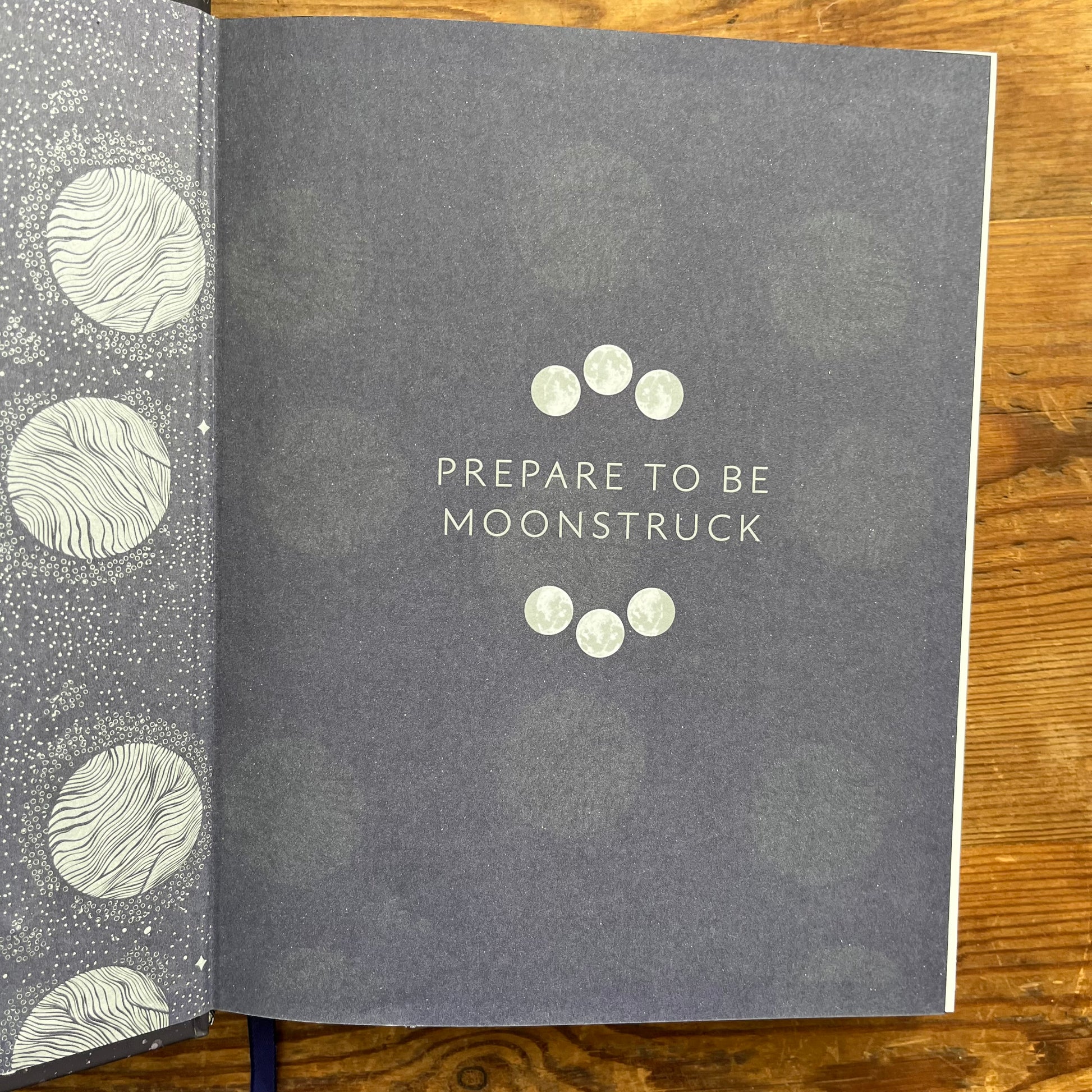 A hardcover journal or book lying open on a wooden surface, showing a dark gray cover with the phrase 'PREPARE TO BE MOONSTRUCK' in white text and moon phases centered on the page. The left page edge has a matching gray-and-white moon pattern.