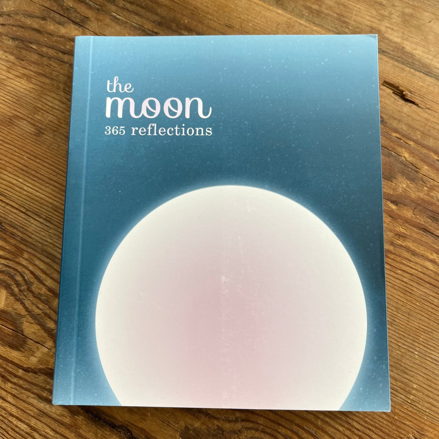 The Moon: 365 Reflections book lying on a wooden surface, featuring a light blue cover with a large white moon graphic and white title text.