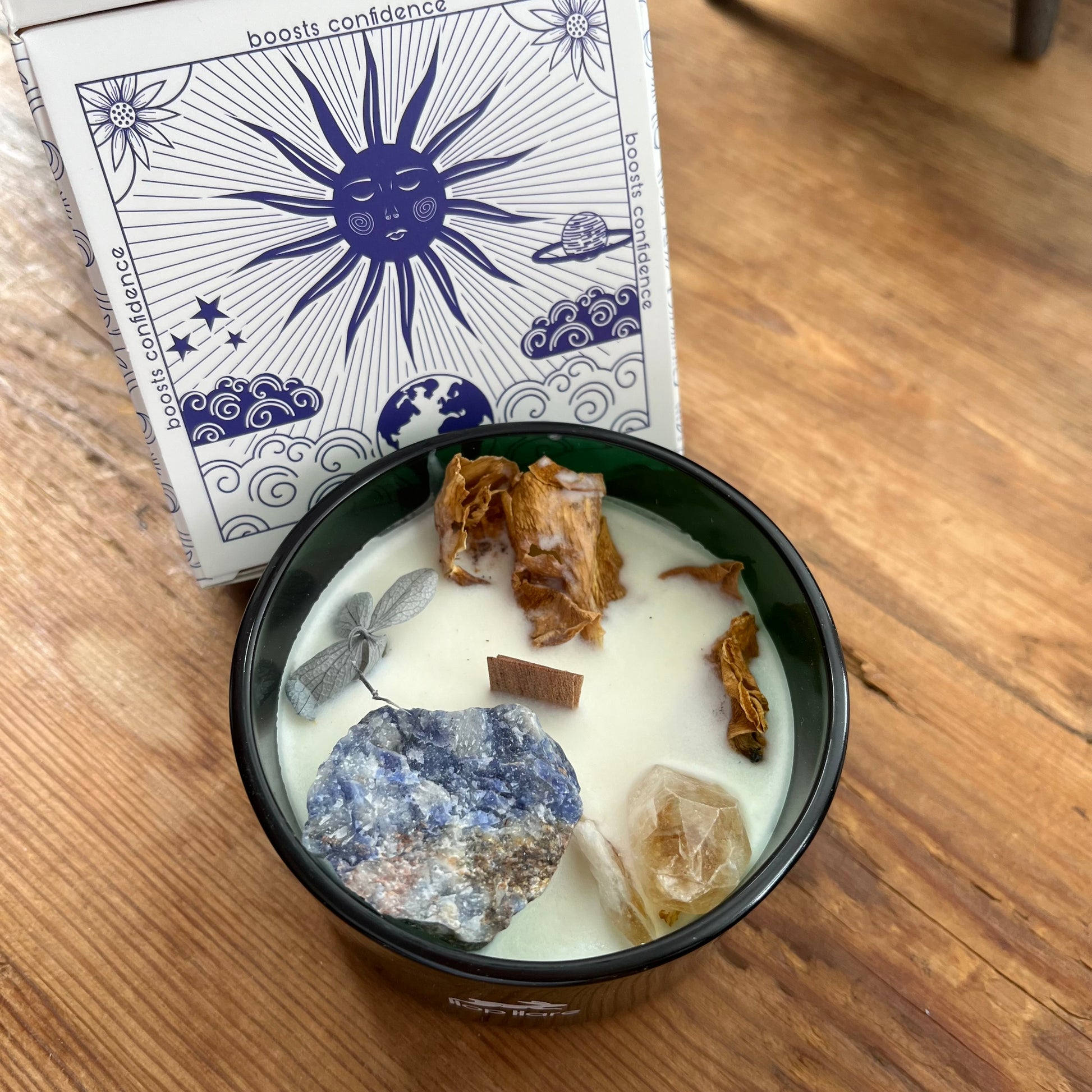 Candle with stones and herbs in a black container on a wooden surface, with a card featuring star and moon designs in the background.