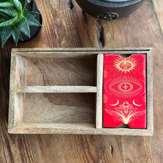 A  wooden divided box suitable for tarot and oracle cards on a rustic wooden table, One compartment has a red lid illustrated with sun and celestrial symbols in gold. with a small succulent in the corner.