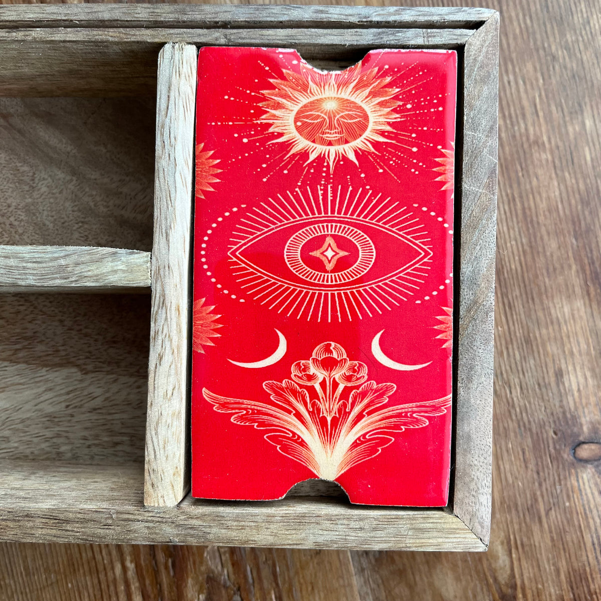 A wooden tarot deck box with ornate gold celestial and floral illustrations, displayed inside a rustic wooden drawer on a wooden tabletop.