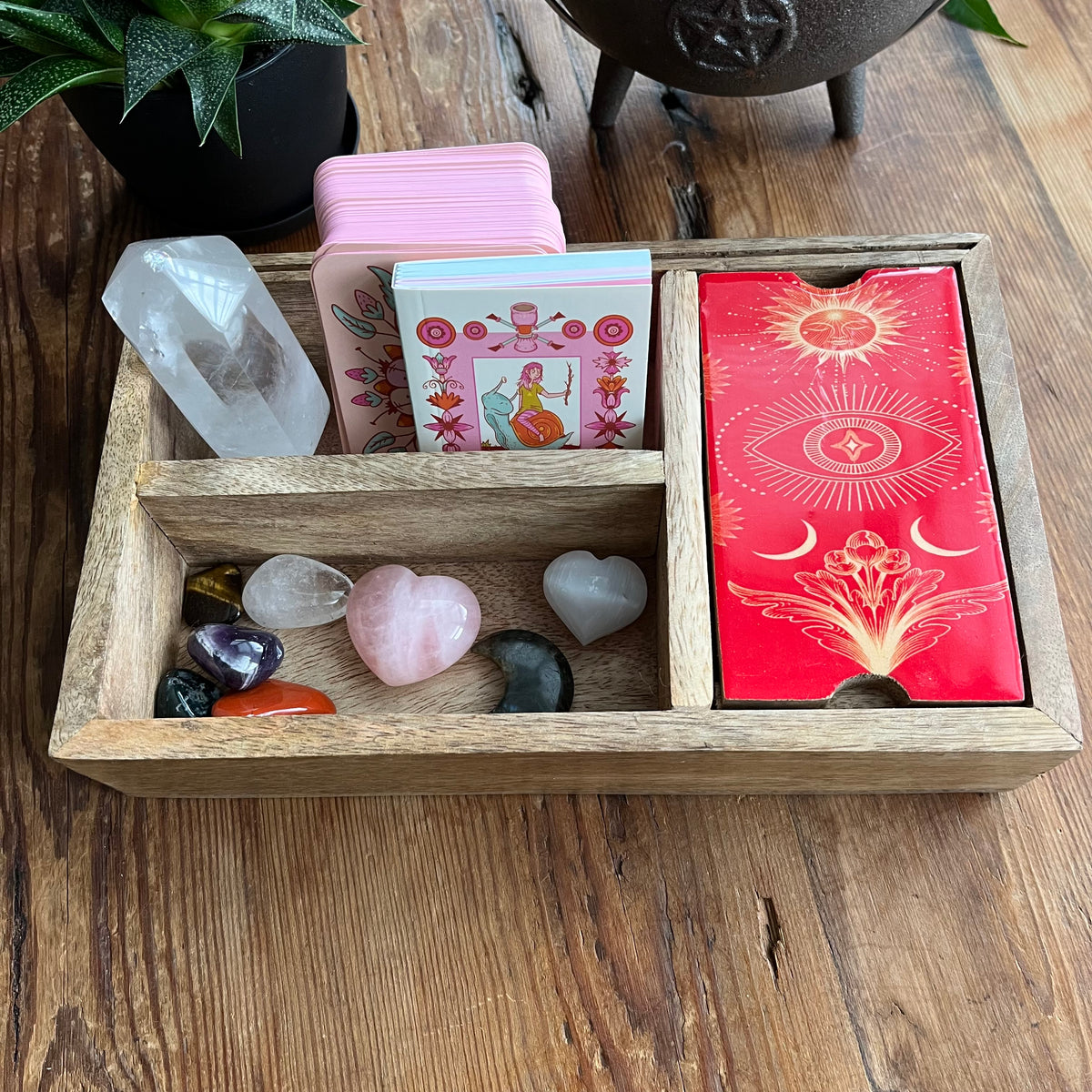 A wooden divided tray holding a pink deck of tarot cards, one compartment has a red lid with gold celestial artwork, another compartment holds a clear crystal point, and several polished stones on a wooden tabletop.