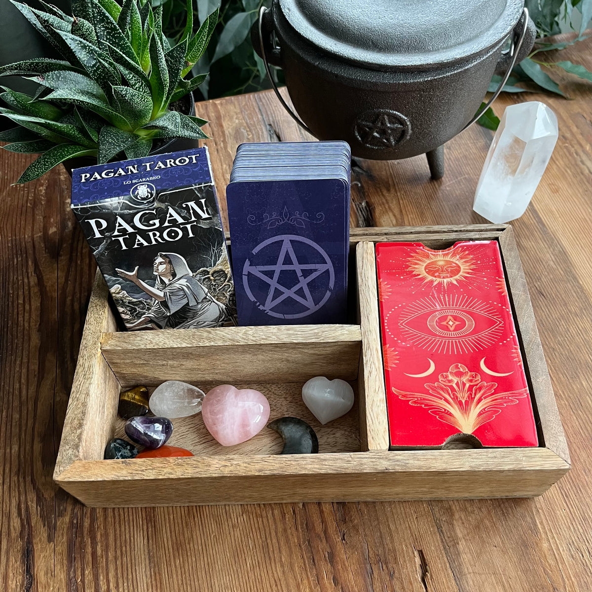 A wooden tray holds a boxed Pagan Tarot deck, a stack of tarot cards with a pentagram design, several crystal stones. it also features a compartment with a red lid with celestial artwork. A black cauldron and green plants sit behind on a wooden table.