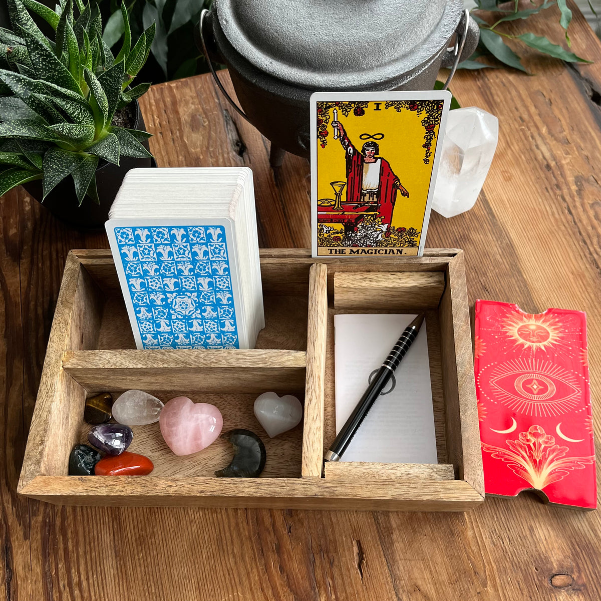 A wooden compartment tray holding a deck of tarot cards, a red lid with gold artwork, a black pen, and several polished crystals on a wooden tabletop. A tarot card reading The Magician is visible, along with a white crystal and a metal container in the background.