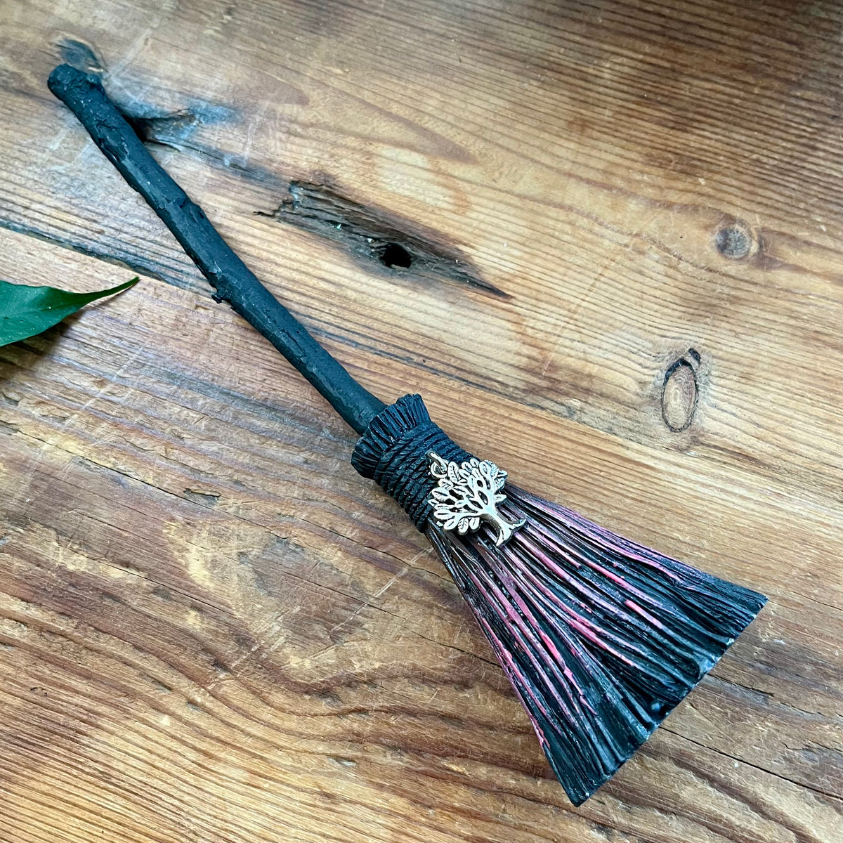 A miniature black witch broomstick with a silver tree ornament and purple-tinted bristles, resting on a rustic wooden surface.