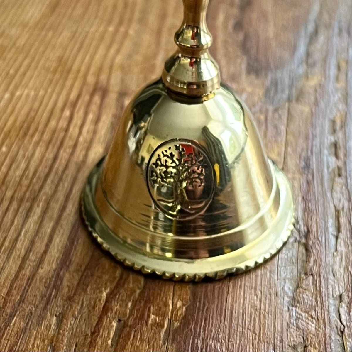 A polished brass bell with a tree of life emblem, resting on a rustic wooden surface.