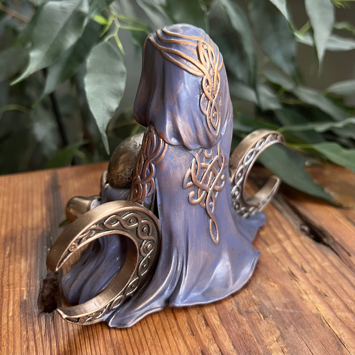 A fantasy-style decorative figurine of a hooded female figure with intricate Celtic knotwork details, displayed on a wooden surface with green foliage in the background.