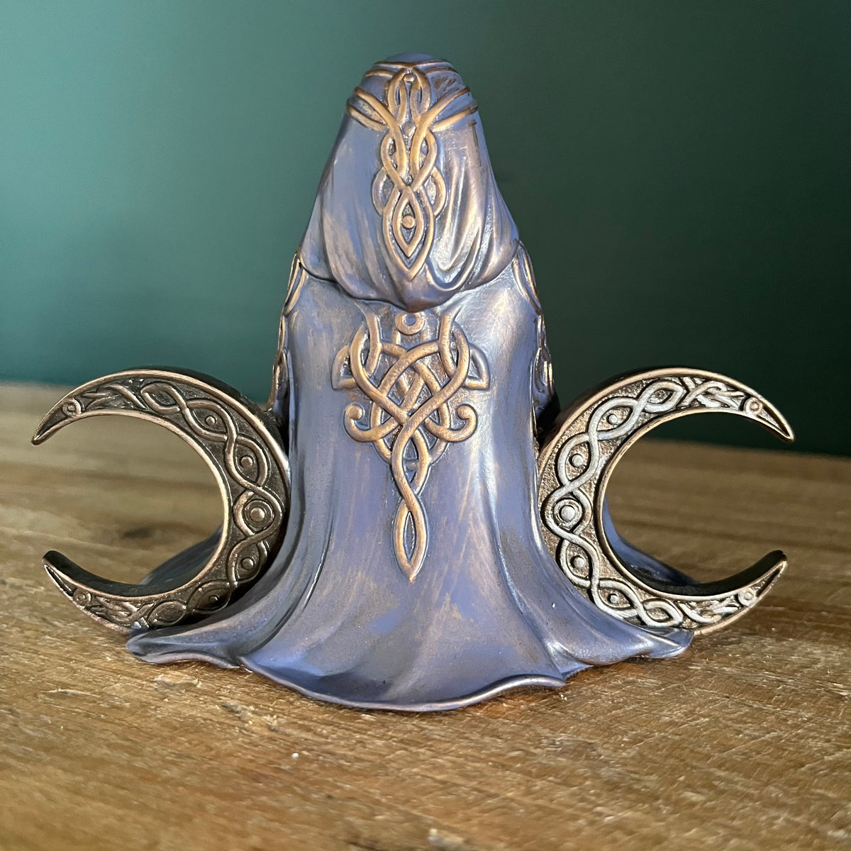 A fantasy-themed metal figurine of a hooded figure with Celtic knotwork engravings, flanked by two crescent moons, displayed on a wooden surface against a dark green background.