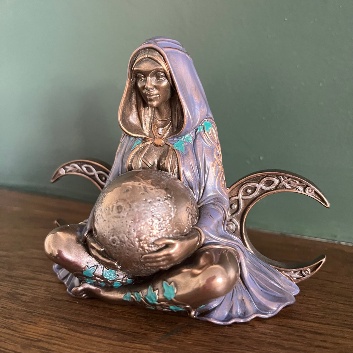 A decorative figurine of a seated hooded woman holding a large metallic sphere, with crescent moon shapes behind her and turquoise accents on a wooden surface against a muted green wall.