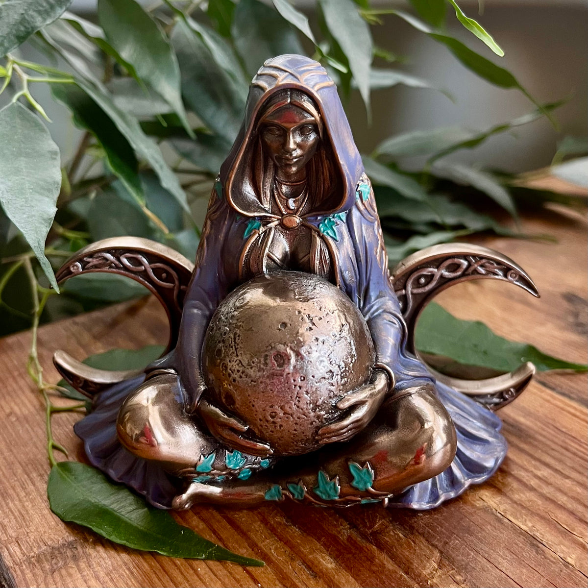 A decorative figurine of a hooded seated woman holding a metallic globe, styled on a wooden surface with green leaves in the background.