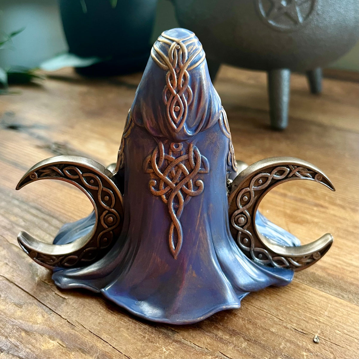 A decorative metal object shaped like a hooded figure with Celtic knotwork details and crescent moon wings, displayed on a wooden surface.