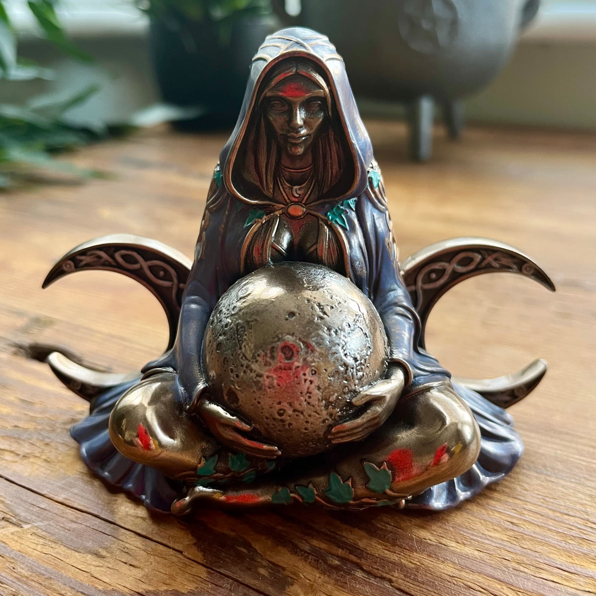 A metal figurine of a hooded figure holding a textured metallic earth orb seated on a wooden surface with decorative crescent-like side elements and small red and green painted accents.