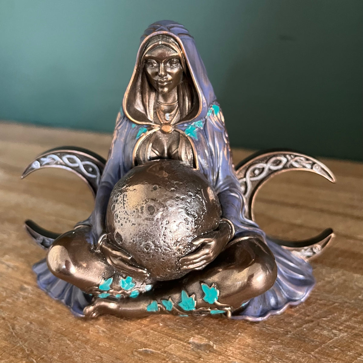 A bronze-finished figurine of a hooded woman seated cross-legged and holding a textured globe, with turquoise accents and crescent moon wings on either side, displayed on a wooden surface against a dark green background.