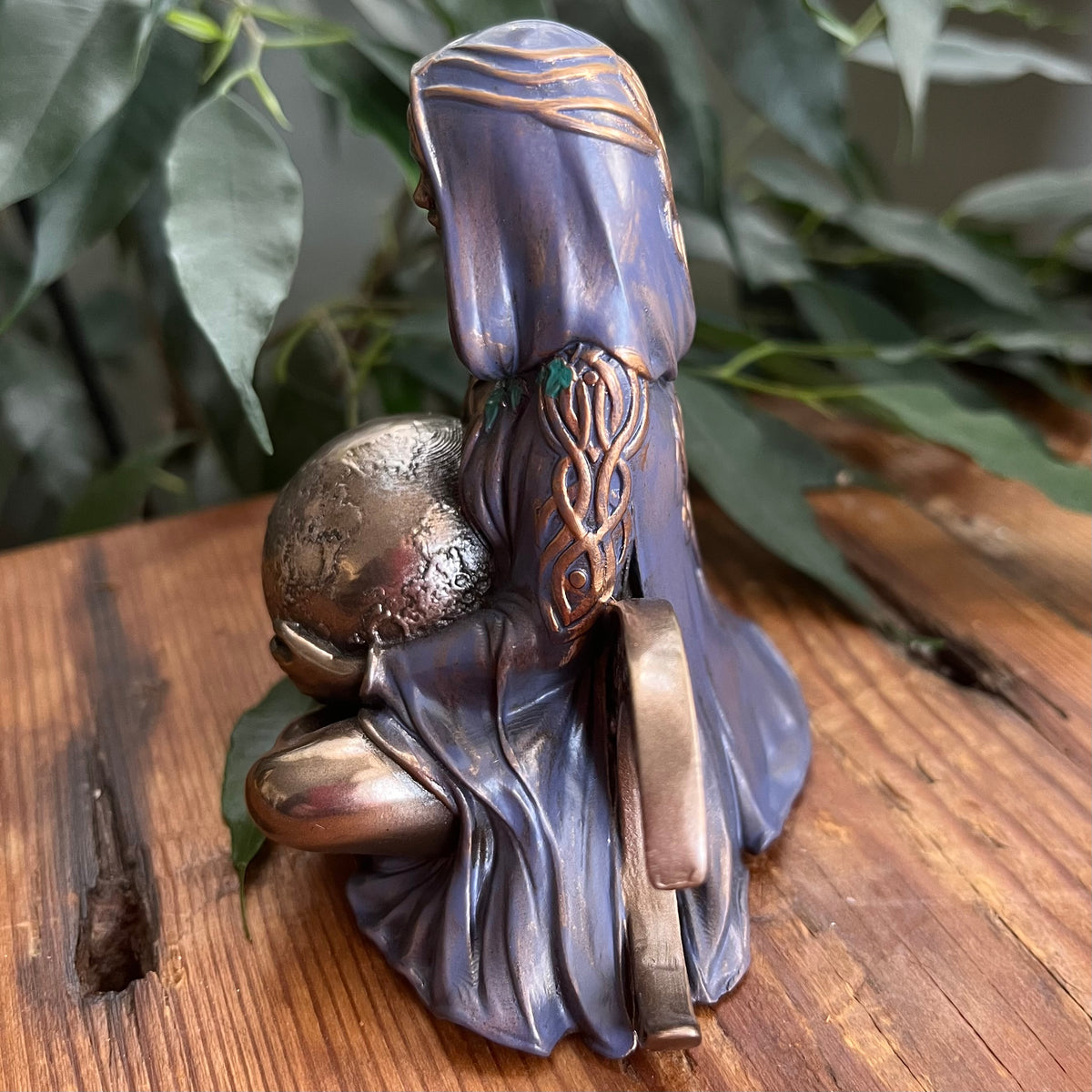 A small figurine of a hooded figure kneeling and holding a textured sphere, shown from the back on a wooden surface with green leaves in the background.