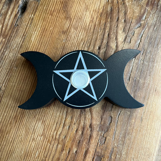 A black triple moon and pentagram spell candle holder resting on a rustic wooden surface.