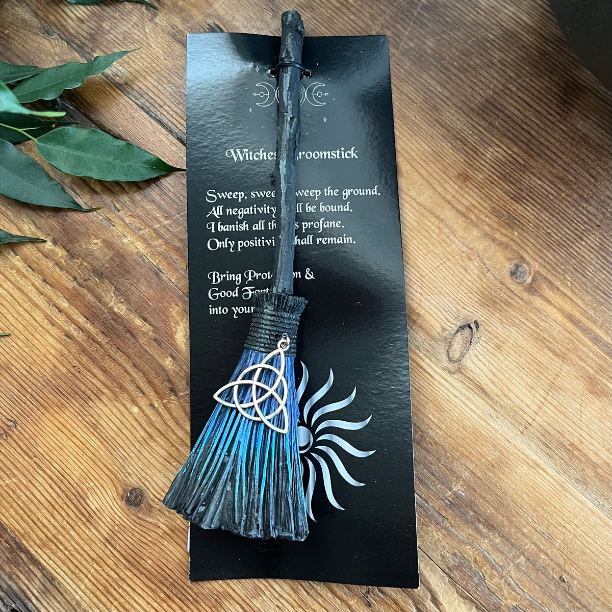 A black wooden witch's broomstick bookmark with a blue and black tassel, featuring a silver Celtic triquetra charm, displayed on its black backing card over a wooden tabletop.