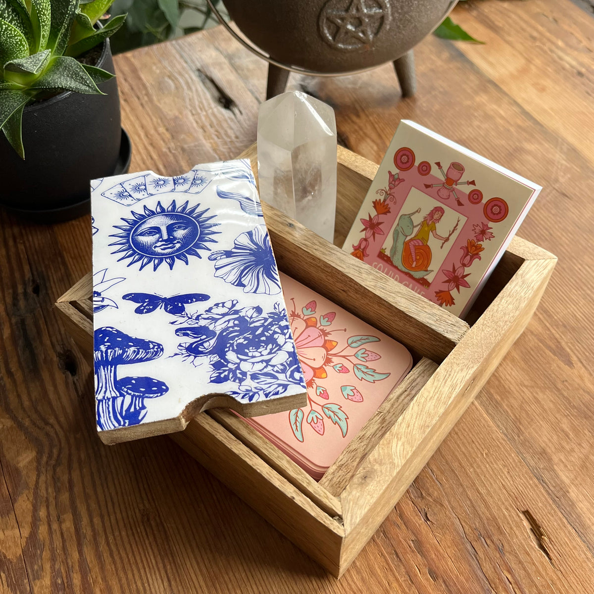 A wooden Tarot or Oracle card storage box holding a stack of decorative cards with a blue-and-white sun-and-floral compartment lid arranged on a wooden table with a crystal and plant nearby.