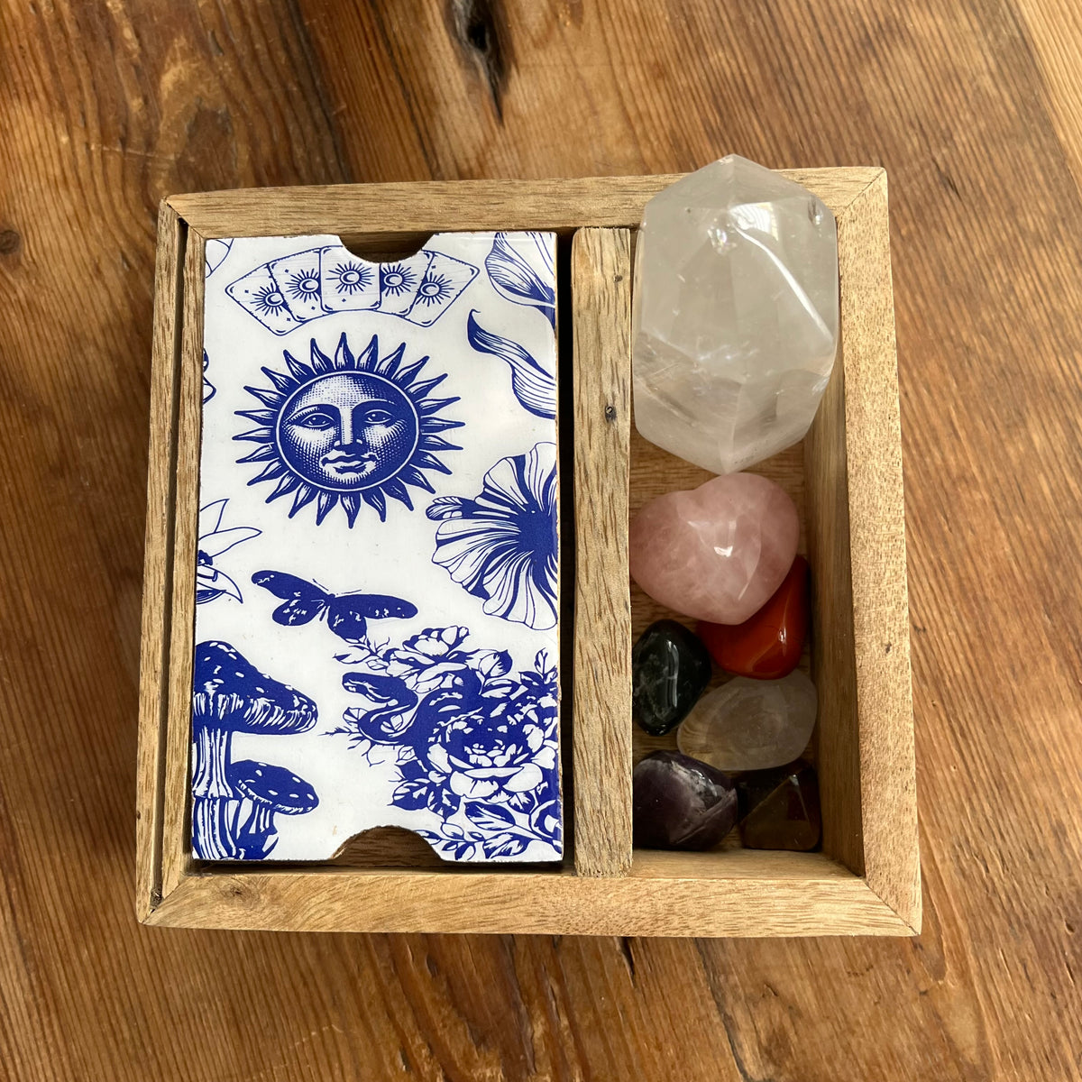 A wooden Tarot or Oracle card storage box with blue illustrated celestial and floral designs, alongside assorted polished crystals on a wooden tabletop.