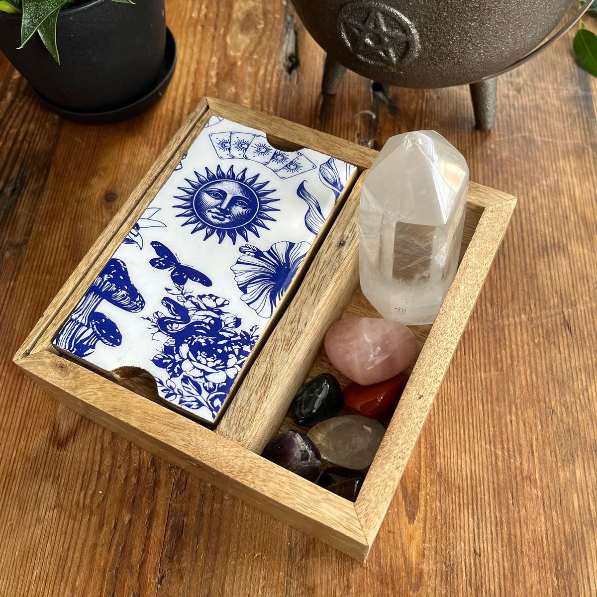 A wooden tray with blue celestial and floral illustrations, alongside a clear crystal tower, pink rose quartz, and polished stones, set on a wooden tabletop.