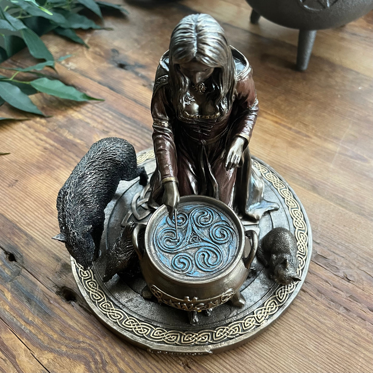 A bronze-finished figurine of a witch beside a cauldron with a boar and other animals on a decorative base, photographed on a wooden tabletop.