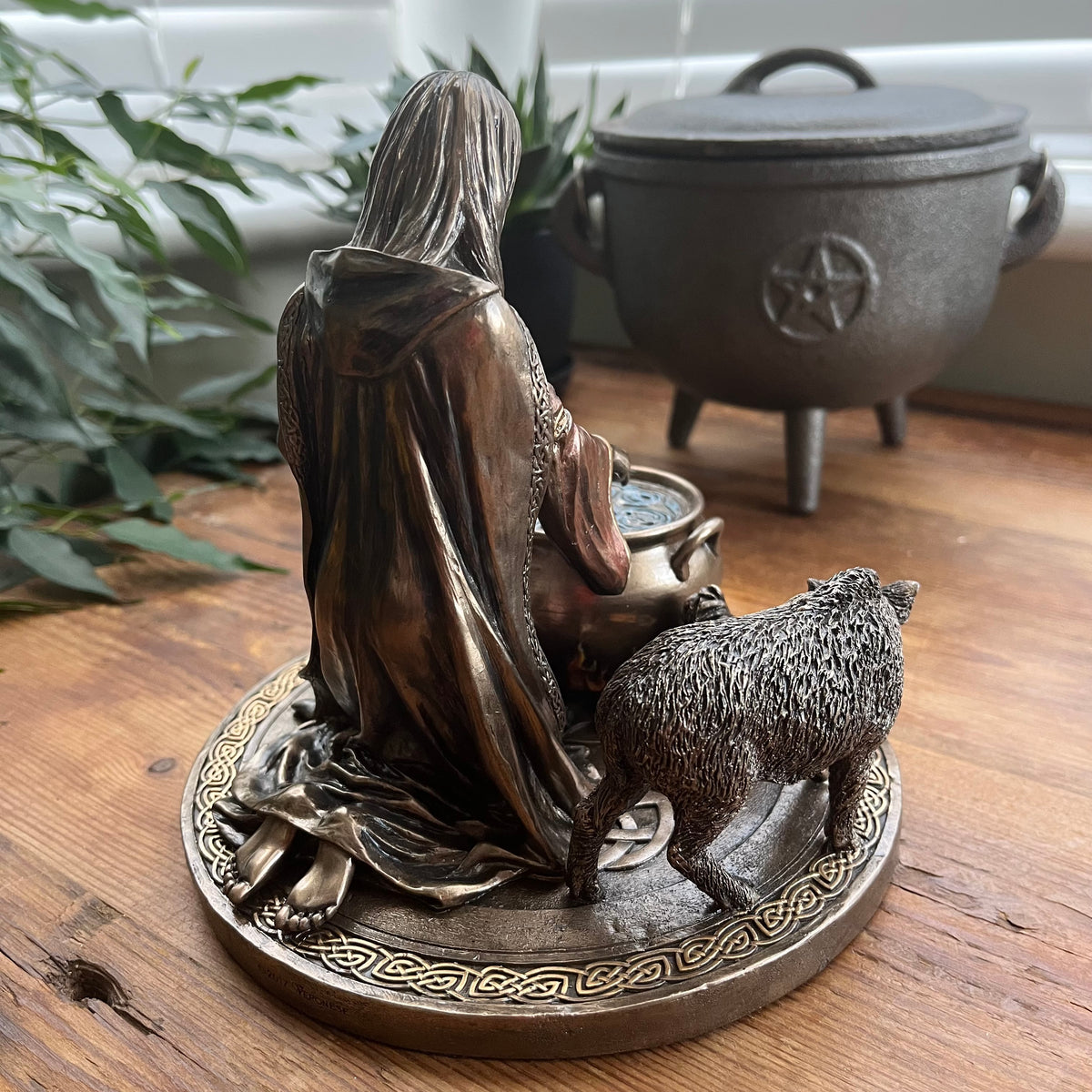 A bronze-finished metal figurine of a hooded figure kneeling beside a cauldron, with a small boar, displayed on a wooden surface with a cauldron and plants in the background.