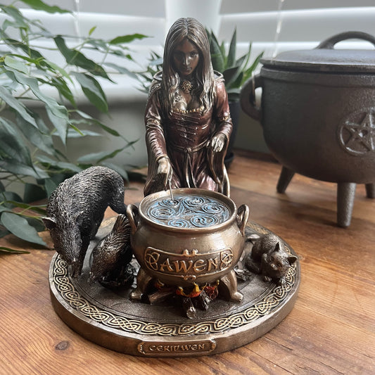 A gothic witch figurine depicting a woman in a brown robe casting a spell over a cauldron labeled 'Awen', accompanied by three Animals on a circular base, displayed on a wooden table.