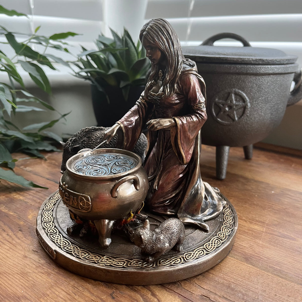 A bronze-finished witch figurine kneeling beside a cauldron with a cat, displayed on a wooden surface with a larger cauldron and green plants in the background.