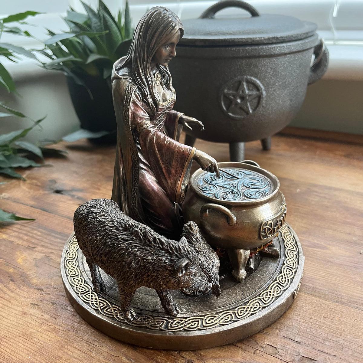 A bronze-finished figurine of a woman kneeling beside a cauldron and a boar displayed on a round decorative base on a wooden surface with a larger cauldron in the blurred background.
