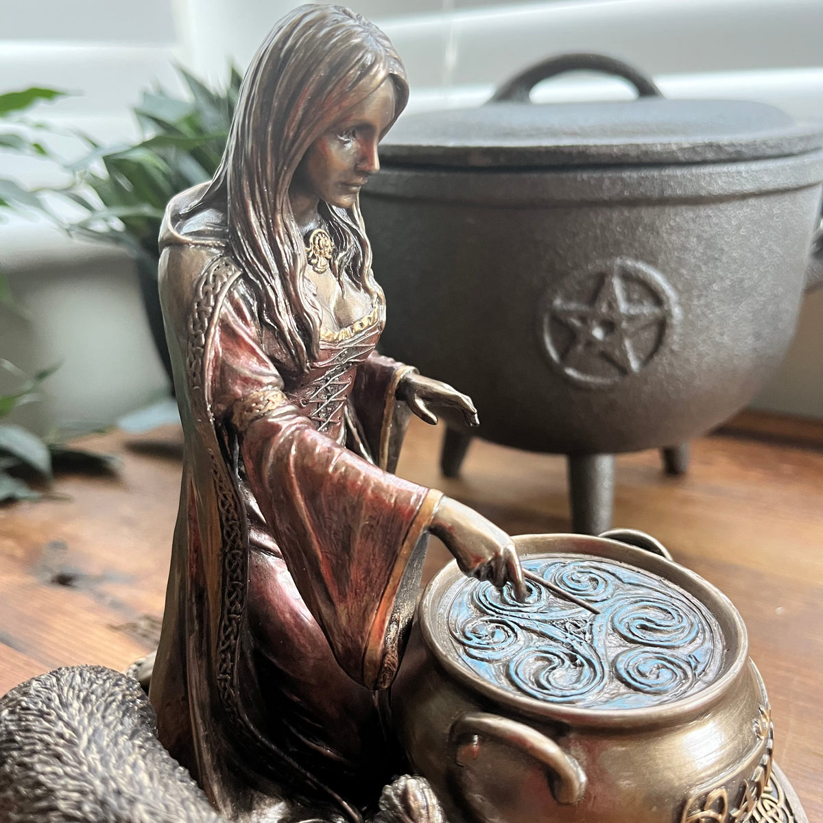 A bronze-finished figurine of a witch kneeling beside a cauldron, with a larger cauldron and a plant blurred in the background on a wooden surface.