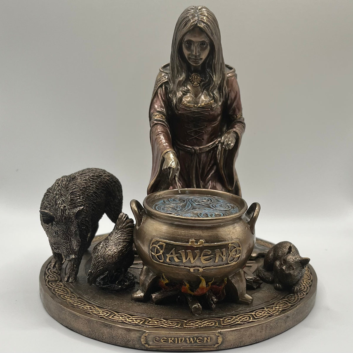 A bronze-finished gothic figurine depicting a witch standing behind a cauldron labeled 'Awen, with a boar, chicken, and cat around it, mounted on a round decorative base.