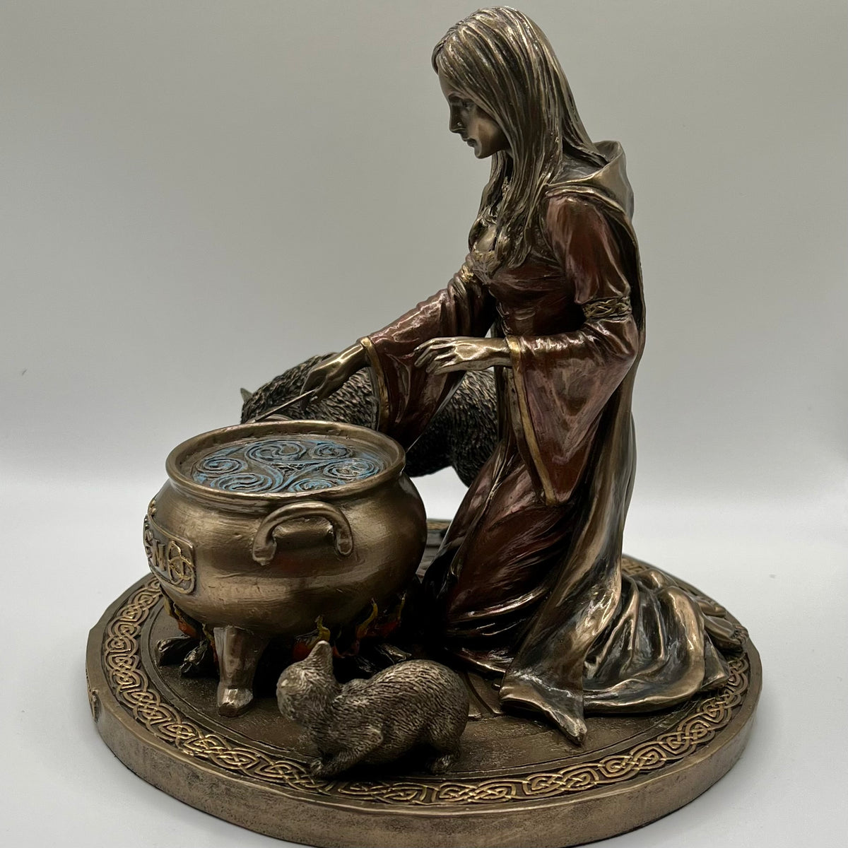 A bronze-finished figurine depicting a kneeling woman beside a cauldron with a small cat in the foreground, displayed on a neutral light gray background.