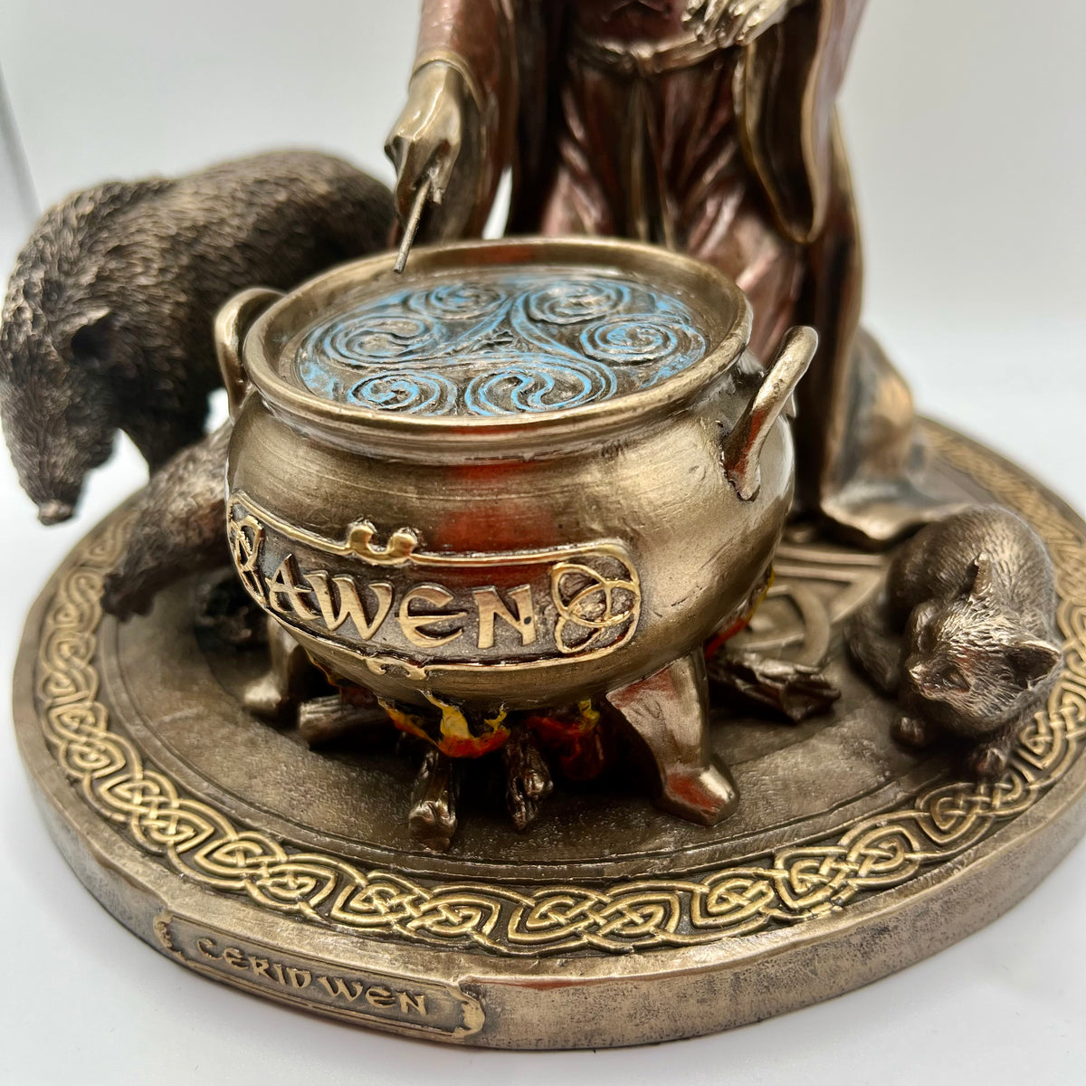 A close-up of a bronze-toned Celtic cauldron figurine with a blue patterned lid, surrounded by animal figures and a circular base engraved with the name 'Awen'.