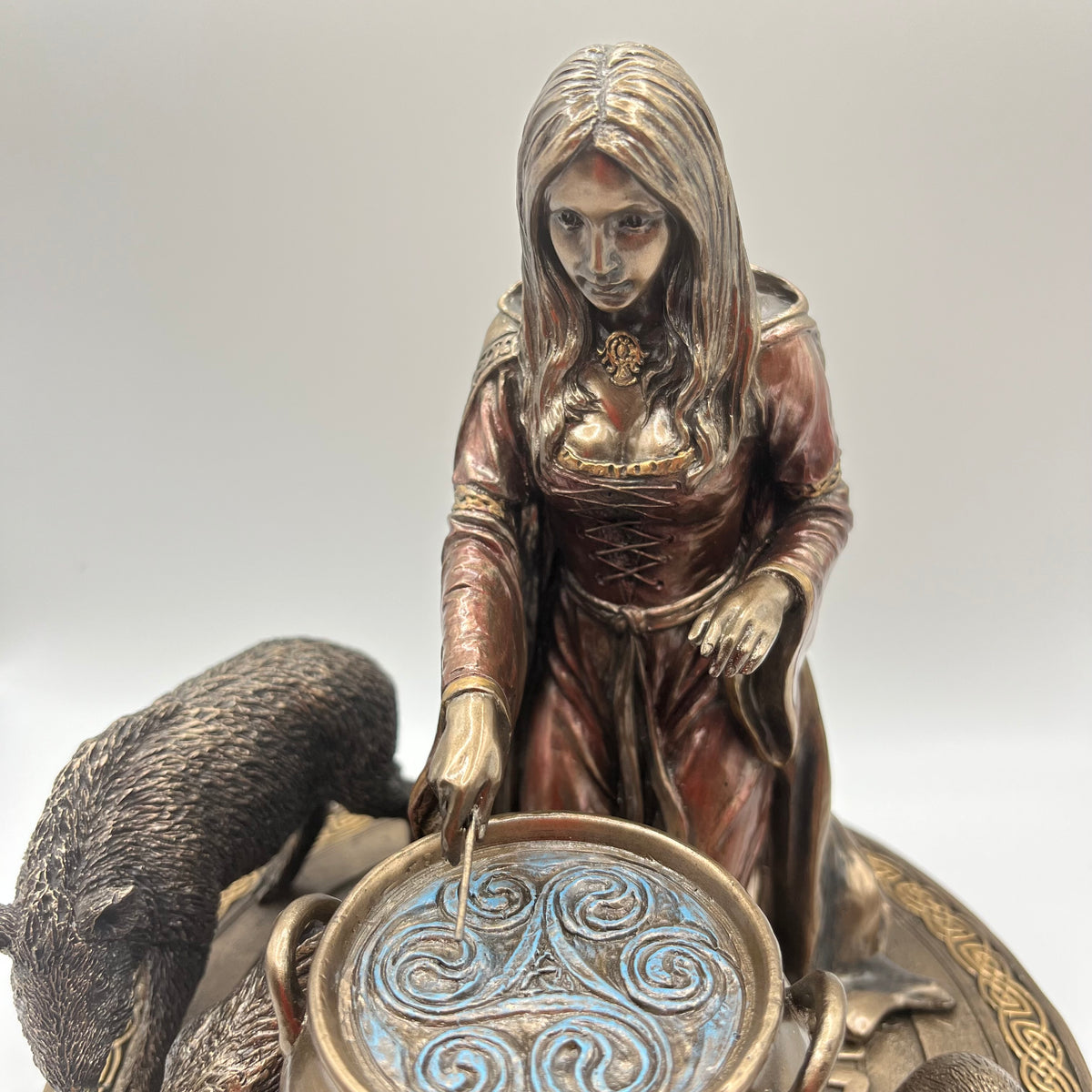A detailed bronze figurine of a woman in a medieval-style dress, leaning over a round decorative bowl with a blue and gold Celtic knot design, accompanied by a dark animal on the left, set against a neutral gray background.