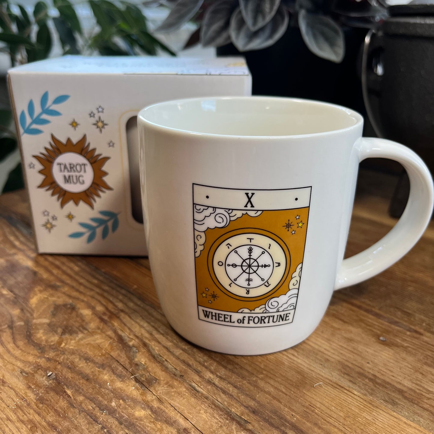 White mug with 'Wheel of Fortune' design on a wooden surface, next to a box labeled 'Tarot Mug'.