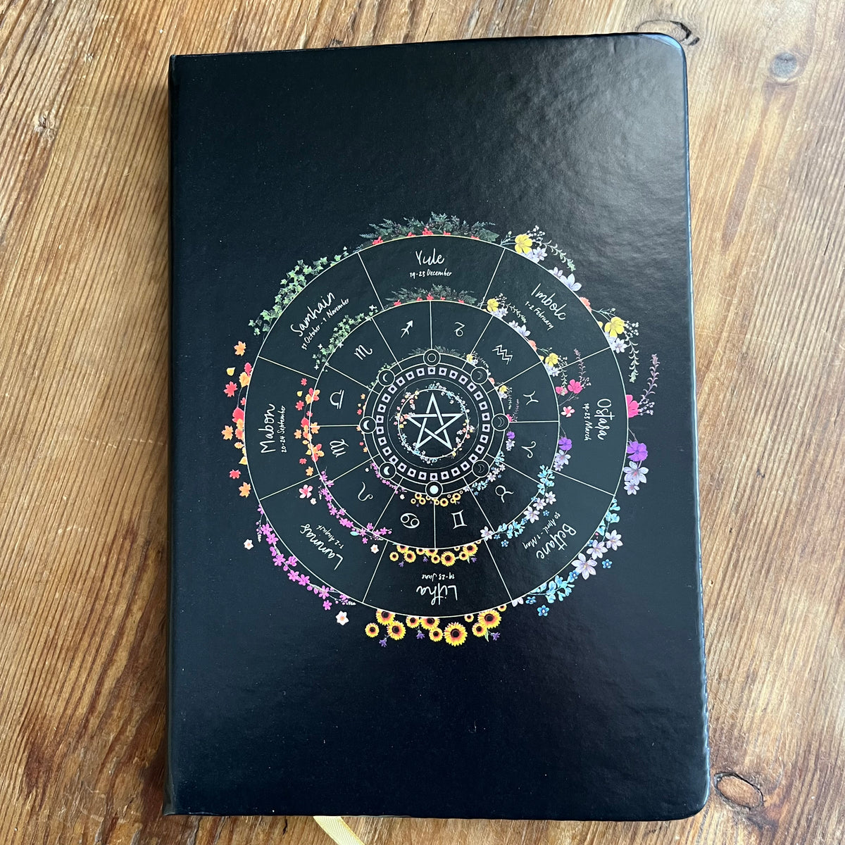 A black hardcover journal featuring a colorful astrological wheel of the year design on the cover, photographed flat on a wooden surface.